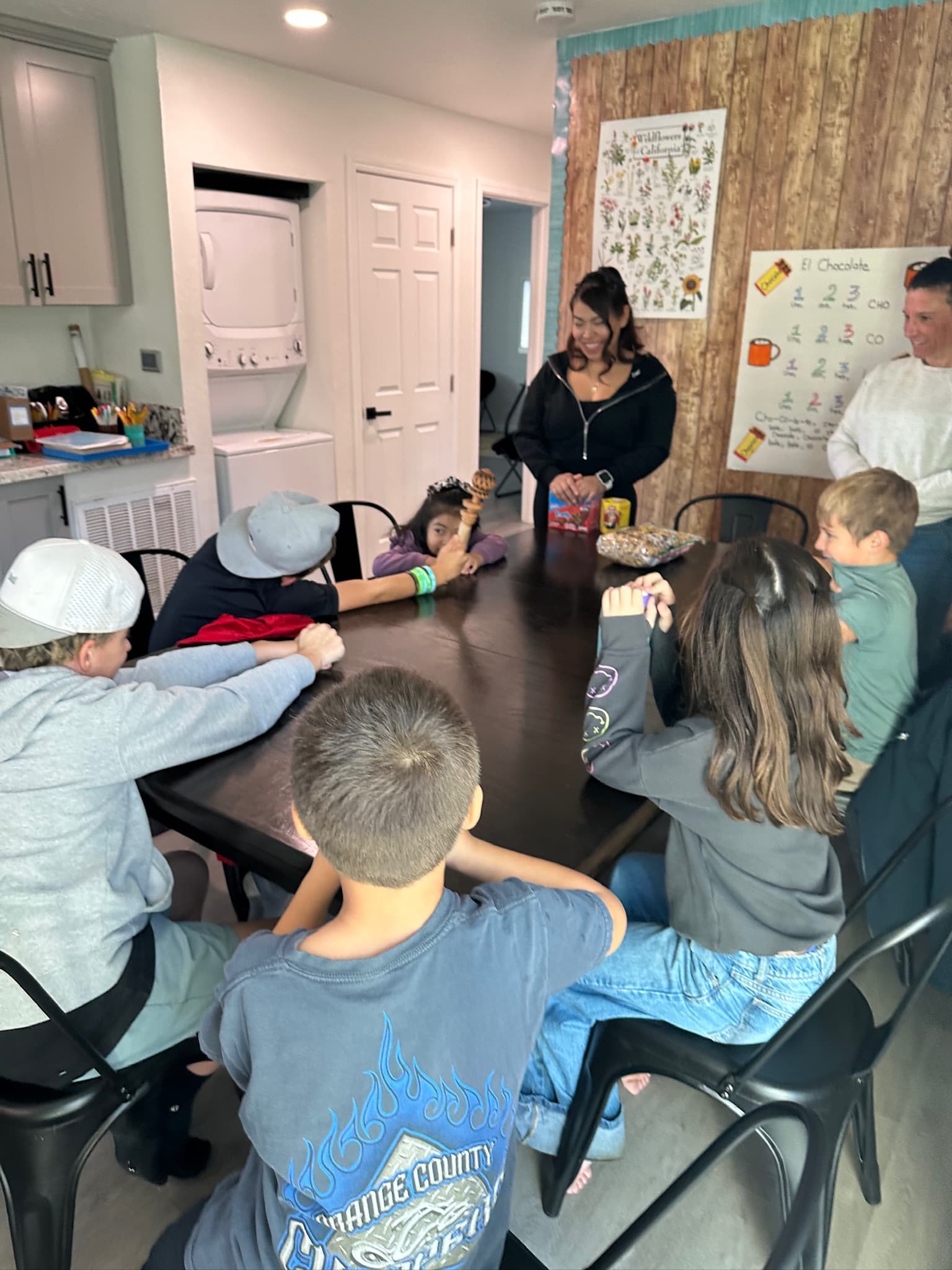Students gathered around a table for a Spanish lesson with a vocabulary chart on the wall
