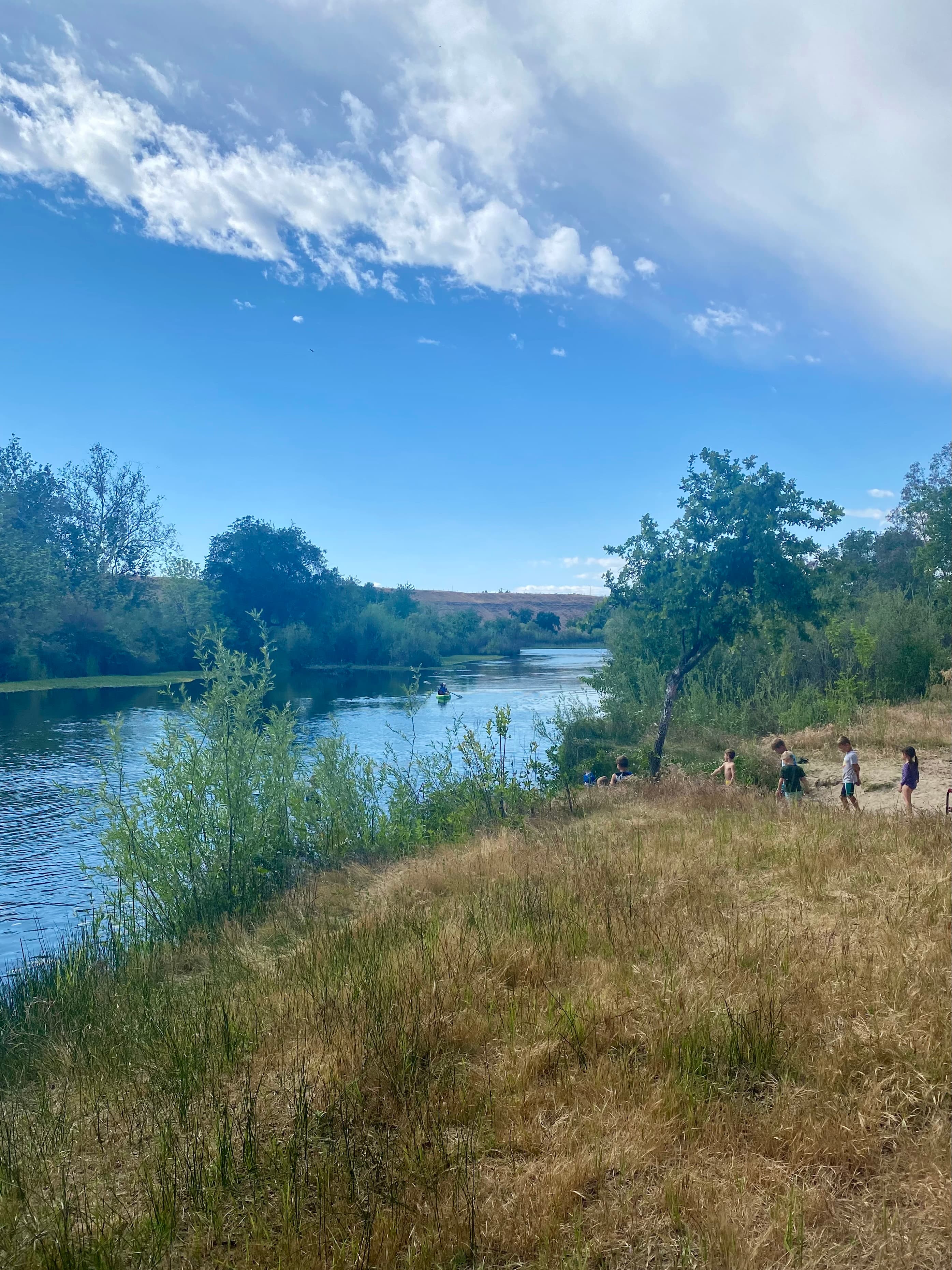 Students walking along the riverbank under a wide blue sky as a kayaker paddles past